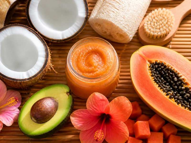 A spa-like arrangement of coconut halves, a jar of sugar scrub, a loofah, and tropical flowers on a bamboo mat.